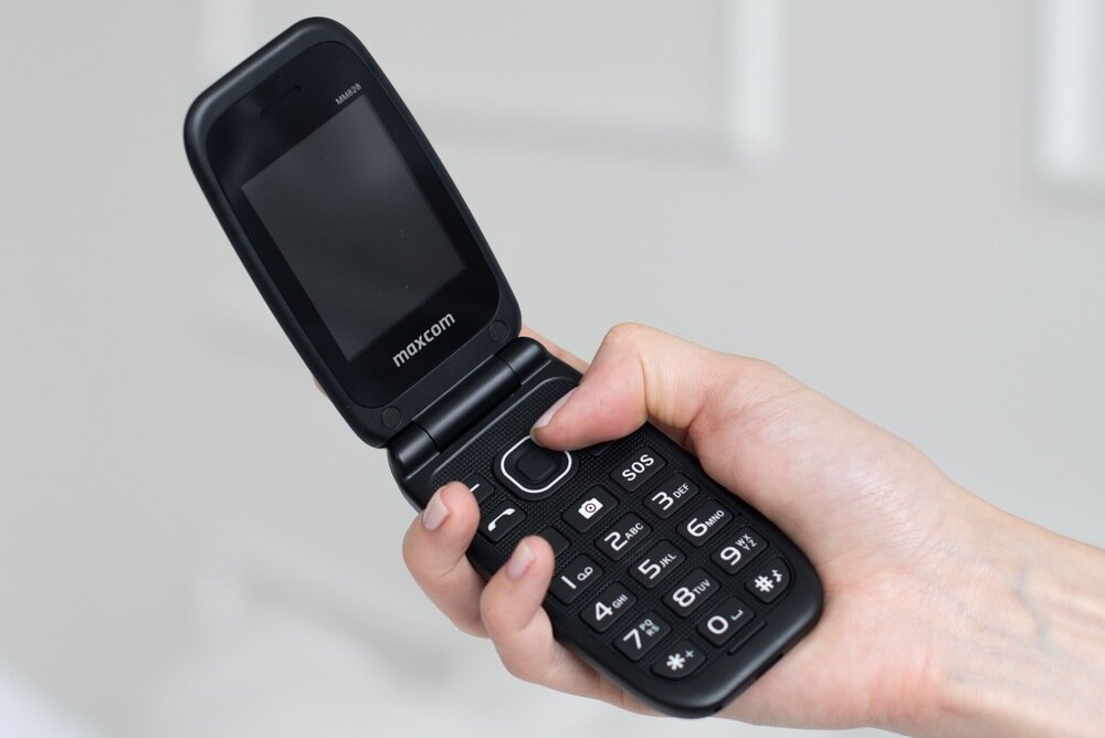 Osoba trzyma otwarty telefon z klapką w kolorze ciemnym. Na ekranie widoczny jest napis \'maxcom\'.