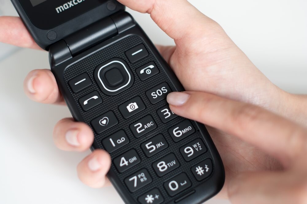 Osoba trzyma telefon z klapką, na którym widoczne są duże przyciski z oznaczeniami, w tym przycisk \'SOS\'. Na górnej części telefonu widnieje logo \'maxcom\'.