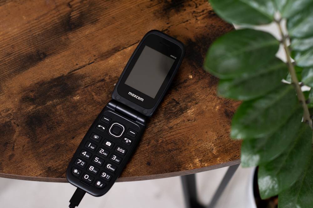 Na drewnianym stole leży otwarty telefon z klapką marki maxcom, z widocznymi dużymi przyciskami i ekranem. Obok znajduje się zielona roślina.