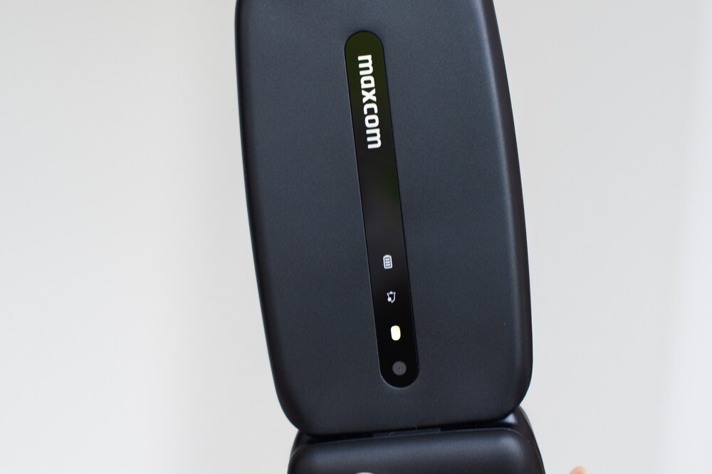 Telefon z klapką w kolorze ciemnym z widocznym logo \'maxcom\' na zewnętrznej stronie. Obok logo znajdują się ikony sygnalizujące stan baterii i połączenia.