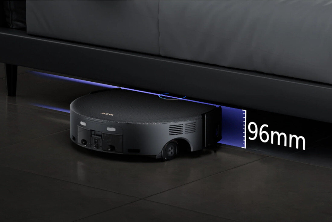 Czarny robot sprzątający znajduje się pod meblem, a obok niego widoczny jest napis \'96mm\'. Na górnej części robota widnieje logo producenta.