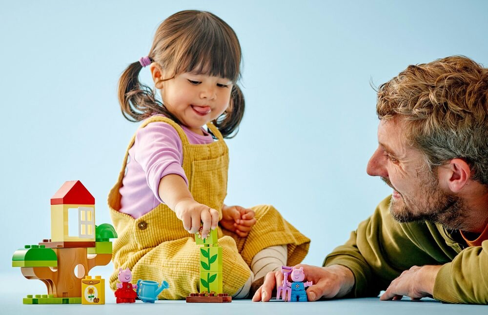 Dziecko i dorosły bawią się klockami LEGO, które przedstawiają kolorowy domek na drzewie i postacie zwierząt. Tło jest jednolite, jasnoniebieskie.