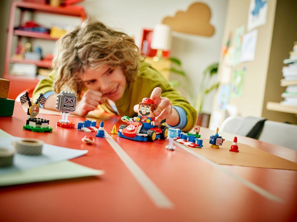 Dziecko bawi się zestawem LEGO, w tym interaktywną figurką Mario i pojazdem. Na stole znajdują się różne elementy toru wyścigowego z klocków LEGO.