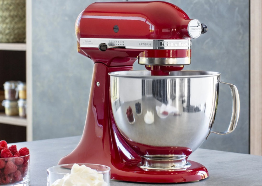 Czerwony robot kuchenny z metalową misą, na której widnieje logo \'KitchenAid\'. W tle znajdują się półki z produktami spożywczymi.