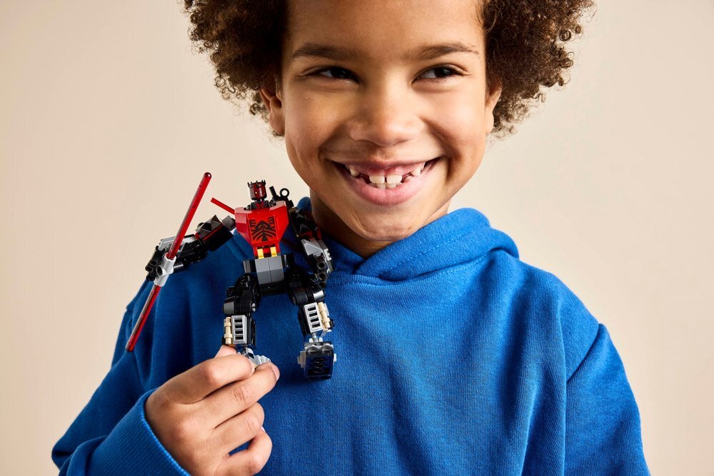 Dziecko w niebieskiej bluzie trzyma figurkę LEGO przedstawiającą mecha z czerwonymi i czarnymi elementami oraz mieczem świetlnym.