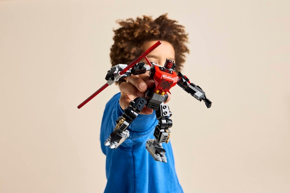 Osoba w niebieskiej bluzie trzyma w ręku figurkę LEGO przedstawiającą robota z czerwonymi i czarnymi elementami oraz mieczem świetlnym.