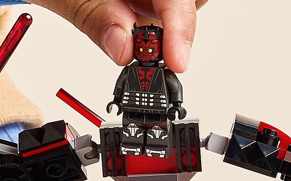 Na zdjęciu widać figurkę LEGO przedstawiającą postać w czarnym stroju z czerwonymi elementami, trzymaną przez rękę. Figurka stoi na konstrukcji z klocków w kolorach czarnym i czerwonym.