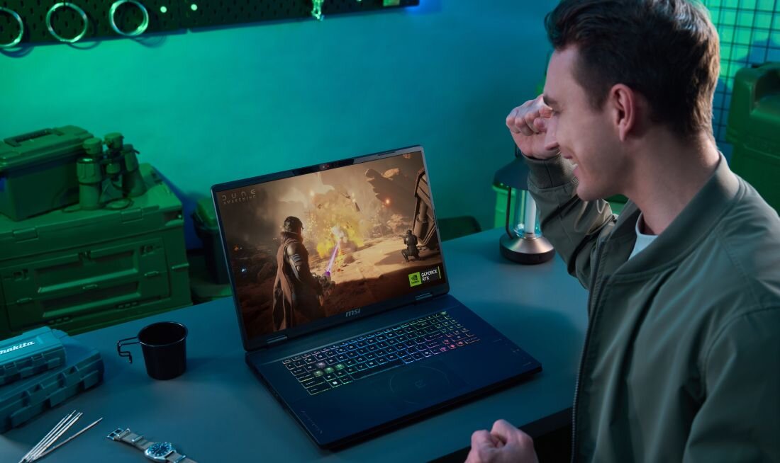 Osoba siedzi przy biurku, na którym znajduje się laptop z wyświetlonym na ekranie dynamicznym obrazem z gry. Na klawiaturze laptopa widoczne jest podświetlenie RGB, a w tle znajdują się różne przedmioty, takie jak kubek i zegarek.