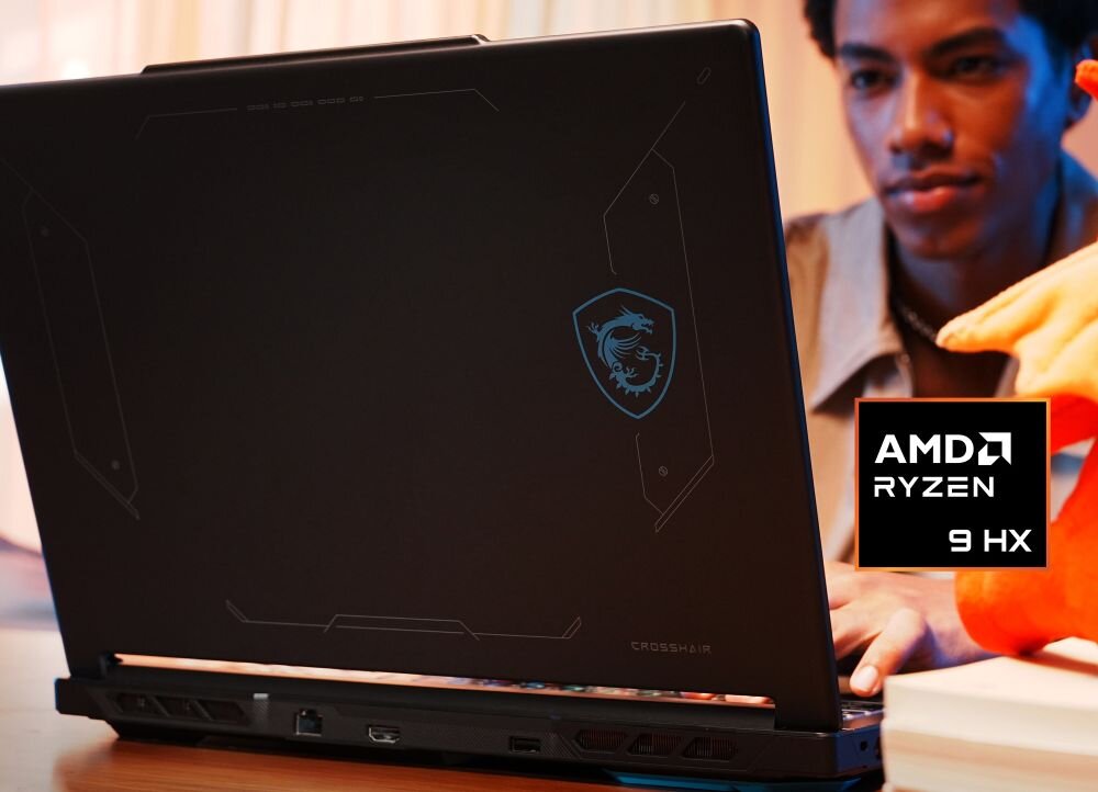 Na zdjęciu widać laptop z logo smoka na obudowie oraz napis \'AMD Ryzen 9 HX\'. W tle znajduje się osoba, a na pierwszym planie widoczny jest fragment pomarańczowego przedmiotu.