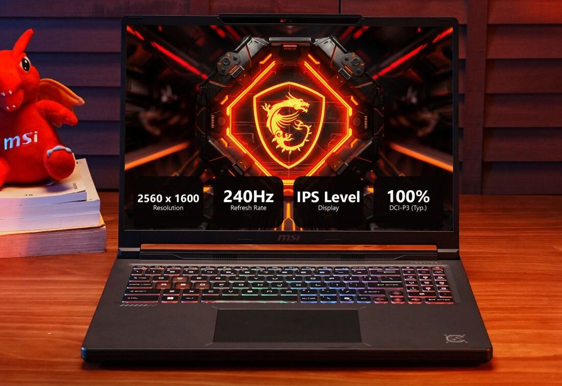 Laptop z wyświetlaczem pokazującym logo smoka i specyfikacje: 2560 x 1600 Resolution, 240Hz Refresh Rate, IPS Level Display, 100% DCI-P3 (Typ.). Obok laptopa znajduje się czerwona maskotka z napisem \'msi\'.