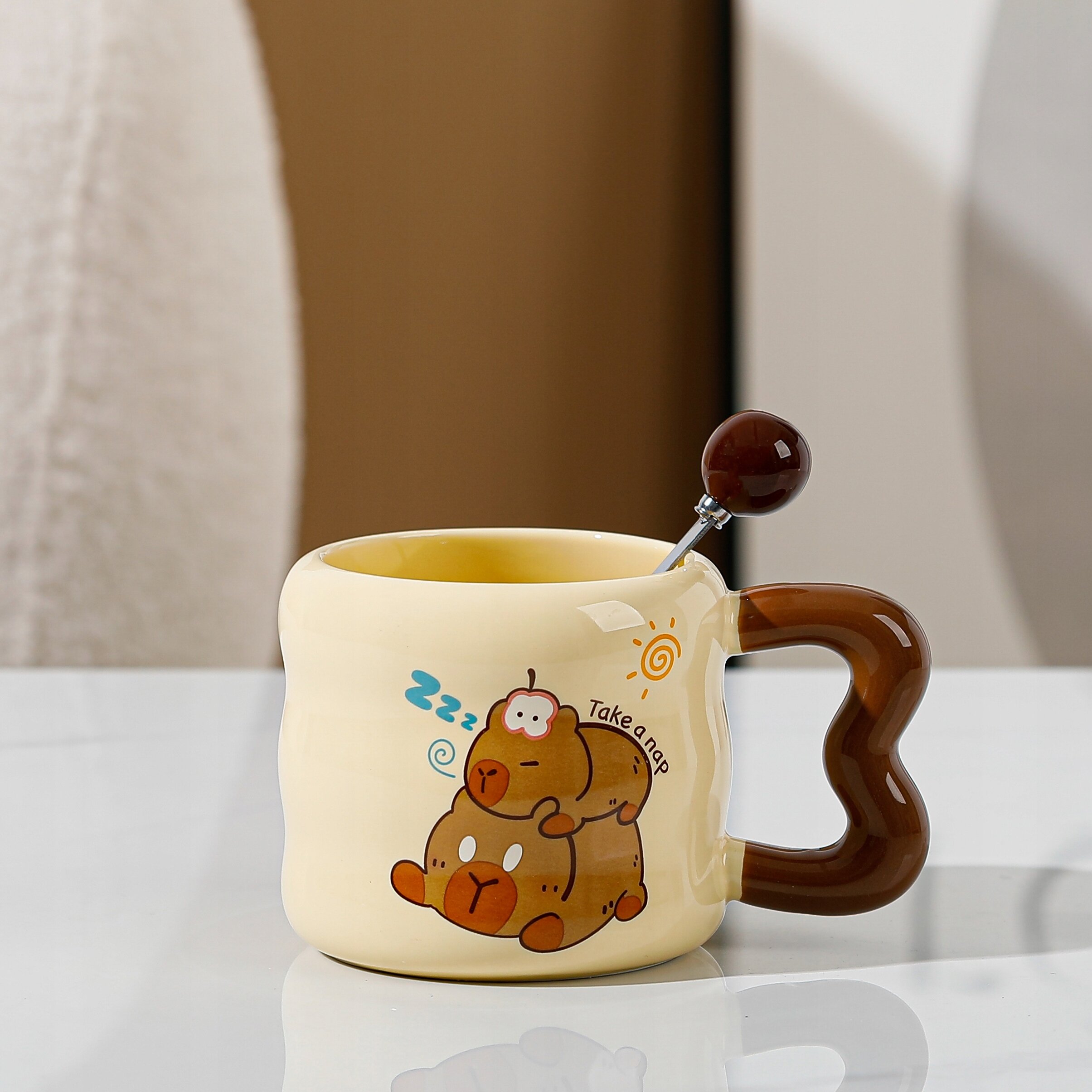Ceramiczny kubek z brązowym uchwytem w kształcie serca, ozdobiony grafiką dwóch śpiących misiów i napisem \'Take a nap\'. W kubku znajduje się łyżeczka z ciemnym uchwytem.