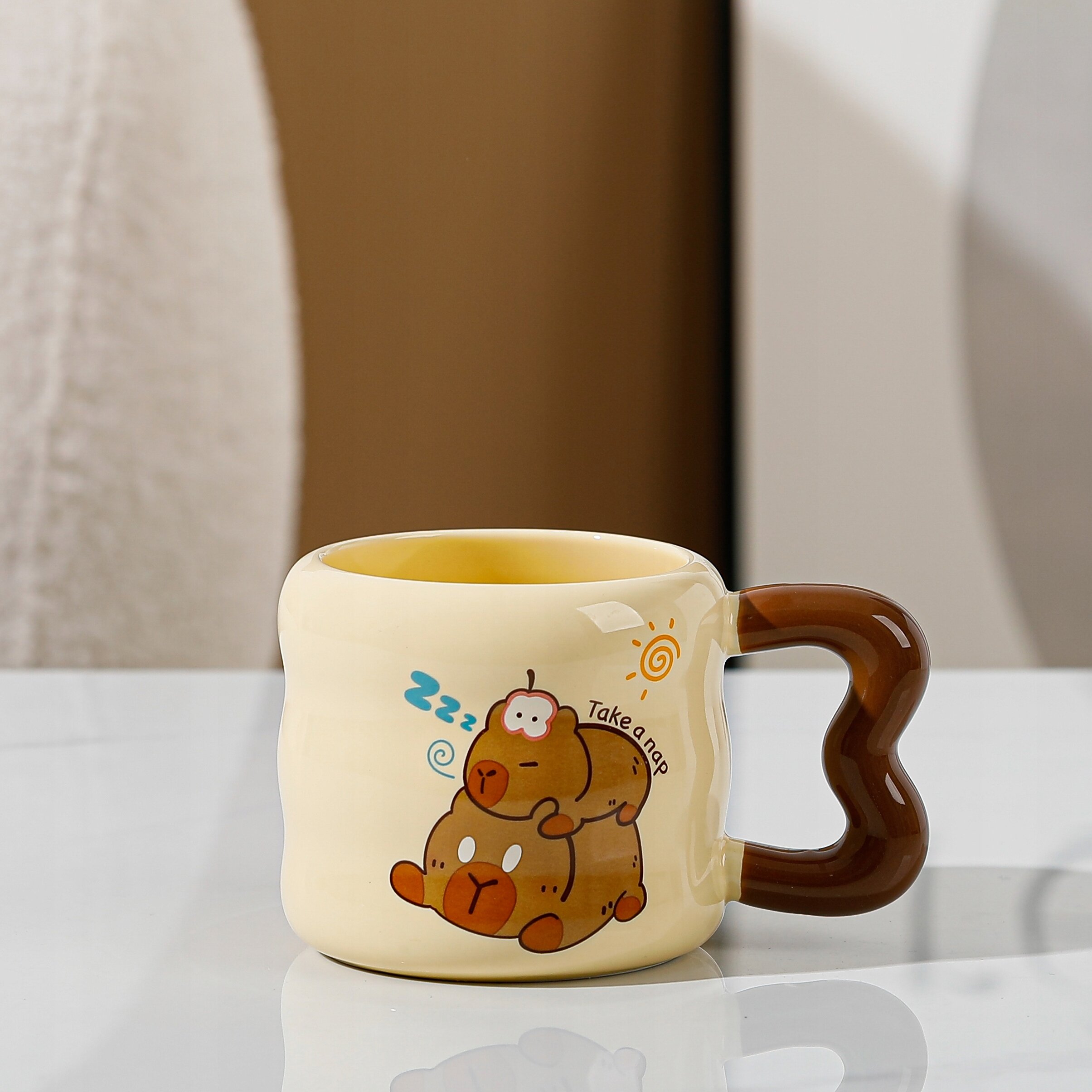 Ceramiczny kubek w jasnym kolorze z brązowym uchwytem w kształcie serca. Na kubku znajduje się ilustracja dwóch śpiących zwierząt z napisem \'Take a nap\'.