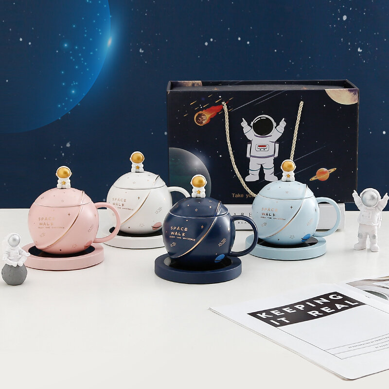 Na obrazie znajdują się cztery ceramiczne kubki w kształcie planet z pokrywkami, każdy w innym kolorze: różowym, białym, granatowym i niebieskim. W tle widoczny jest karton z grafiką astronauty i planet, a na stole leży otwarta książka z napisem \'KEEPING IT REAL\'.