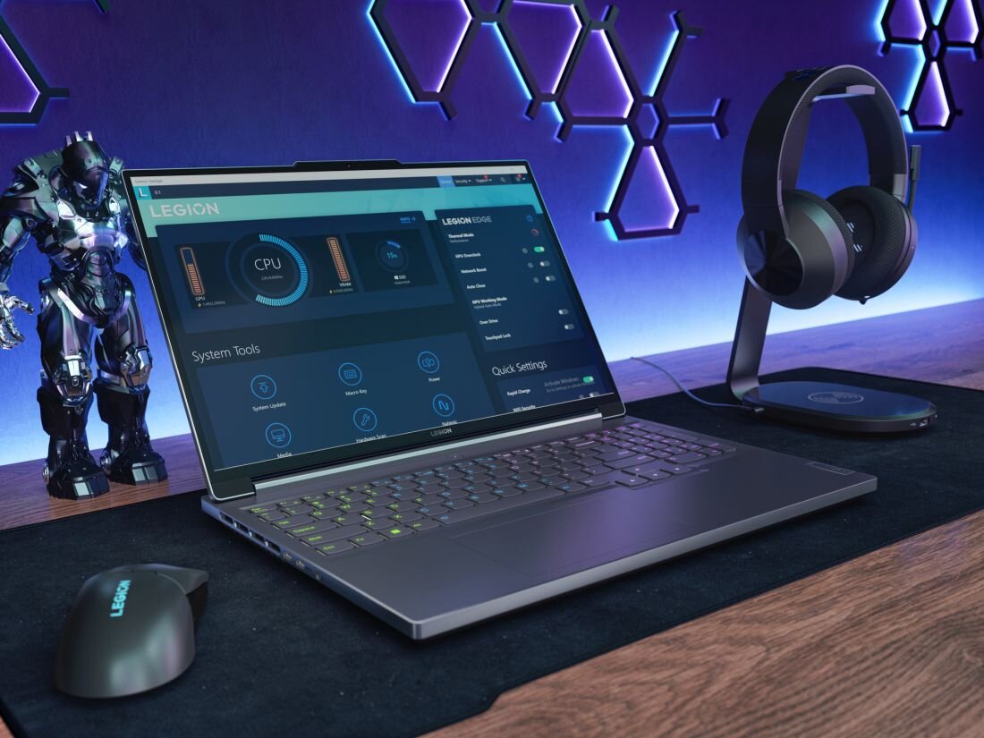 Na biurku znajduje się laptop z włączonym ekranem, obok myszka z napisem \'LEGION\' i stojak na słuchawki. W tle widoczna jest figurka robota oraz ściana z geometrycznymi wzorami podświetlonymi na niebiesko.