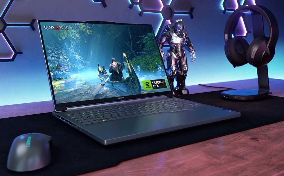 Na biurku znajduje się laptop z wyświetlonym na ekranie obrazem z gry \'God of War\' oraz logo \'GEFORCE GTX\'. Obok laptopa widoczne są słuchawki na stojaku, myszka i figurka postaci w zbroi.