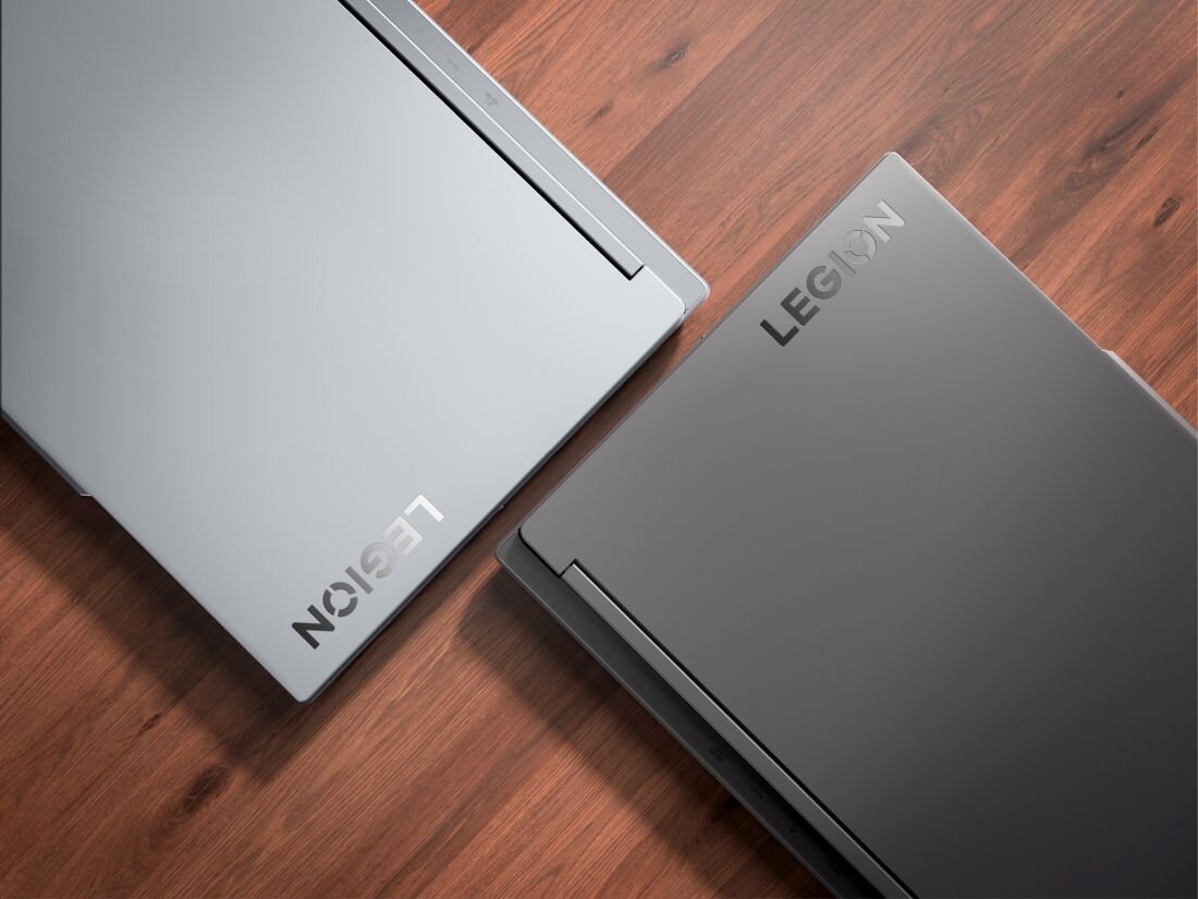 Na drewnianej powierzchni znajdują się dwa zamknięte laptopy z widocznym napisem \'LEGION\' na obudowach. Jeden laptop jest jasnoszary, a drugi ciemnoszary.