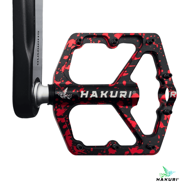 Czarno-czerwony pedał rowerowy z pinami, z napisem \'HAKURI\' i logo. Widoczna jest również część ramienia korby z napisem \'Shimano\'.