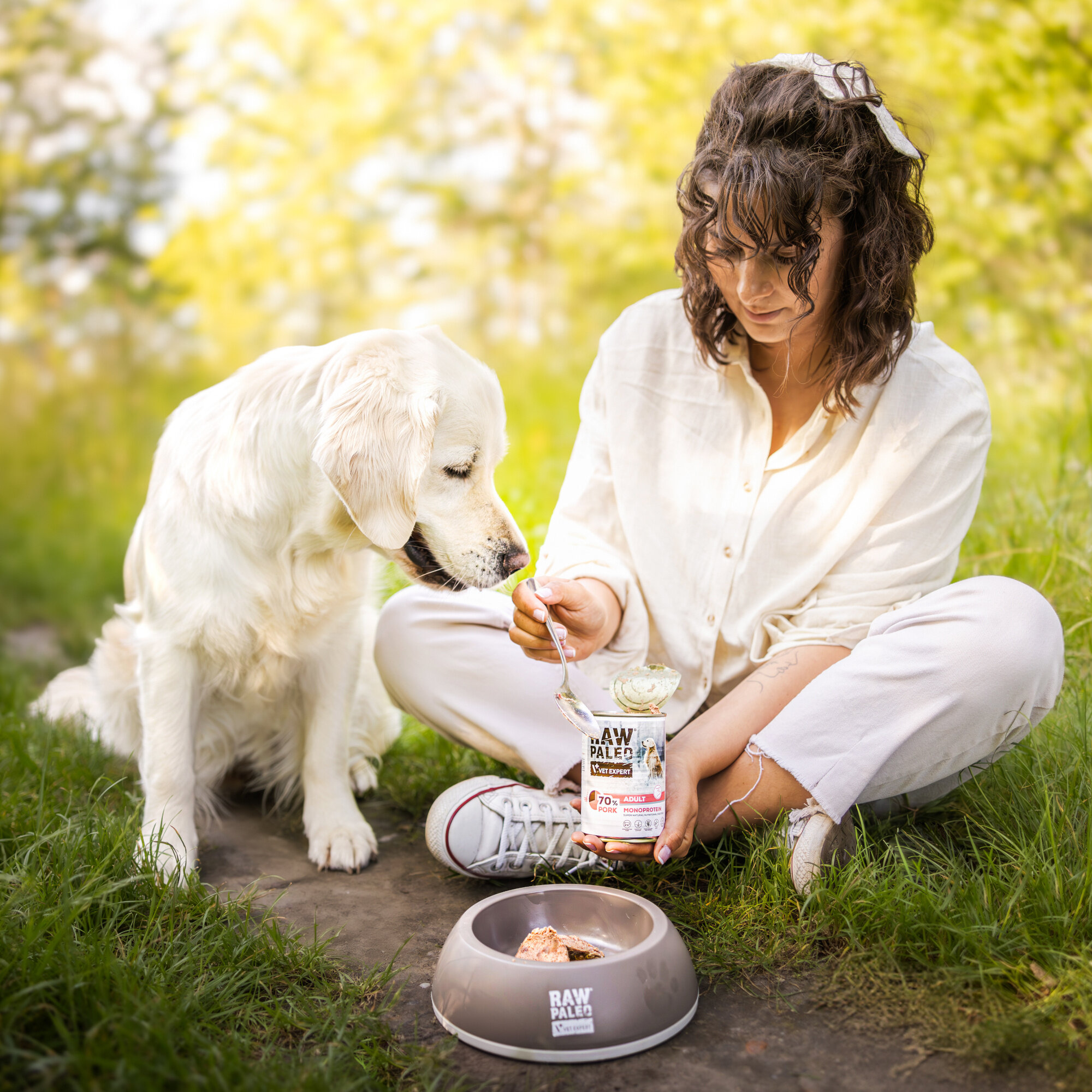 Osoba siedzi na trawie obok psa, trzymając puszkę karmy RAW PALEO ADULT DOG PORK 400g. Przed nimi znajduje się miska z karmą, a w tle widać rozmyte drzewa.