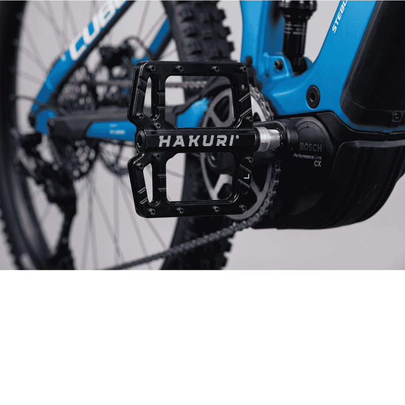 Na zdjęciu widać czarny, aluminiowy pedał rowerowy z napisem \'HAKURI\' zamontowany na niebieskim rowerze. Obok pedału widoczne jest logo \'BOSCH Performance Line CX\'.