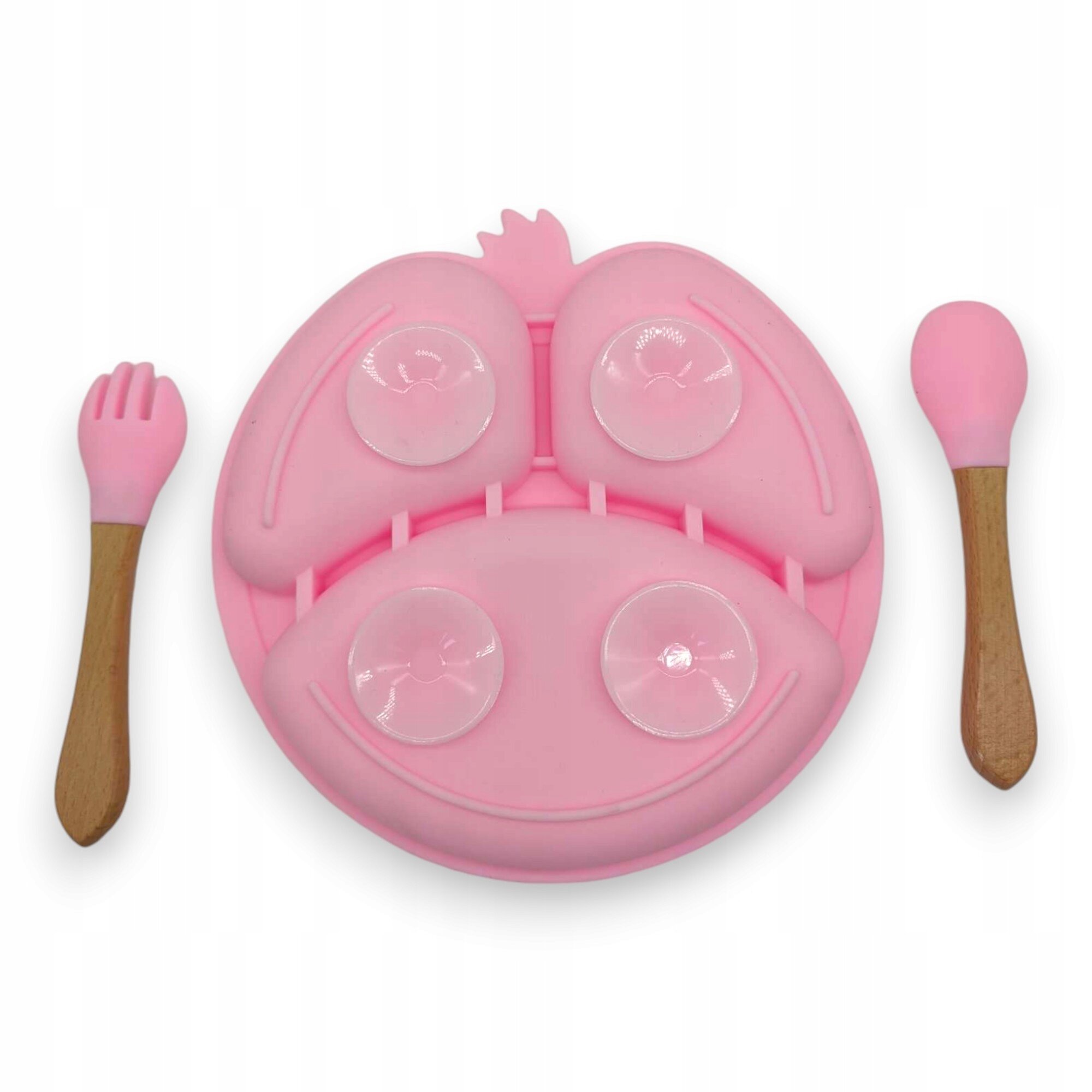 Różowy silikonowy talerzyk z przyssawkami w kształcie uśmiechniętej twarzy, obok którego znajdują się różowe sztućce z drewnianymi uchwytami.
