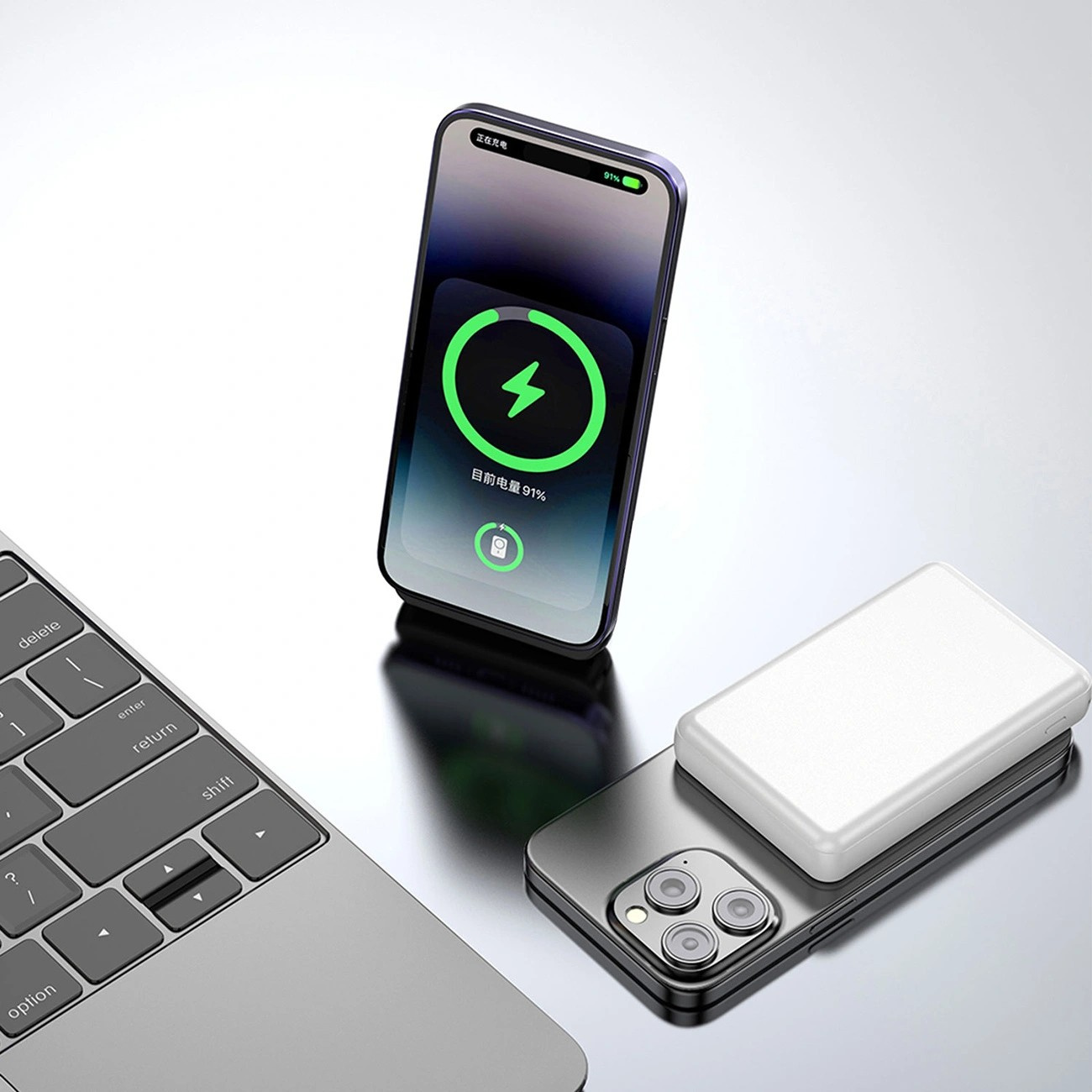 Na obrazie widoczny jest telefon z wyświetlonym symbolem ładowania oraz biały powerbank leżący na innym telefonie. Obok znajduje się fragment klawiatury laptopa.