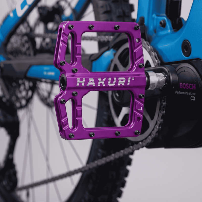 Fioletowy pedał rowerowy z napisem \'HAKURI\' zamontowany na rowerze. W tle widoczna jest część roweru z napędem \'BOSCH Performance Line CX\'.