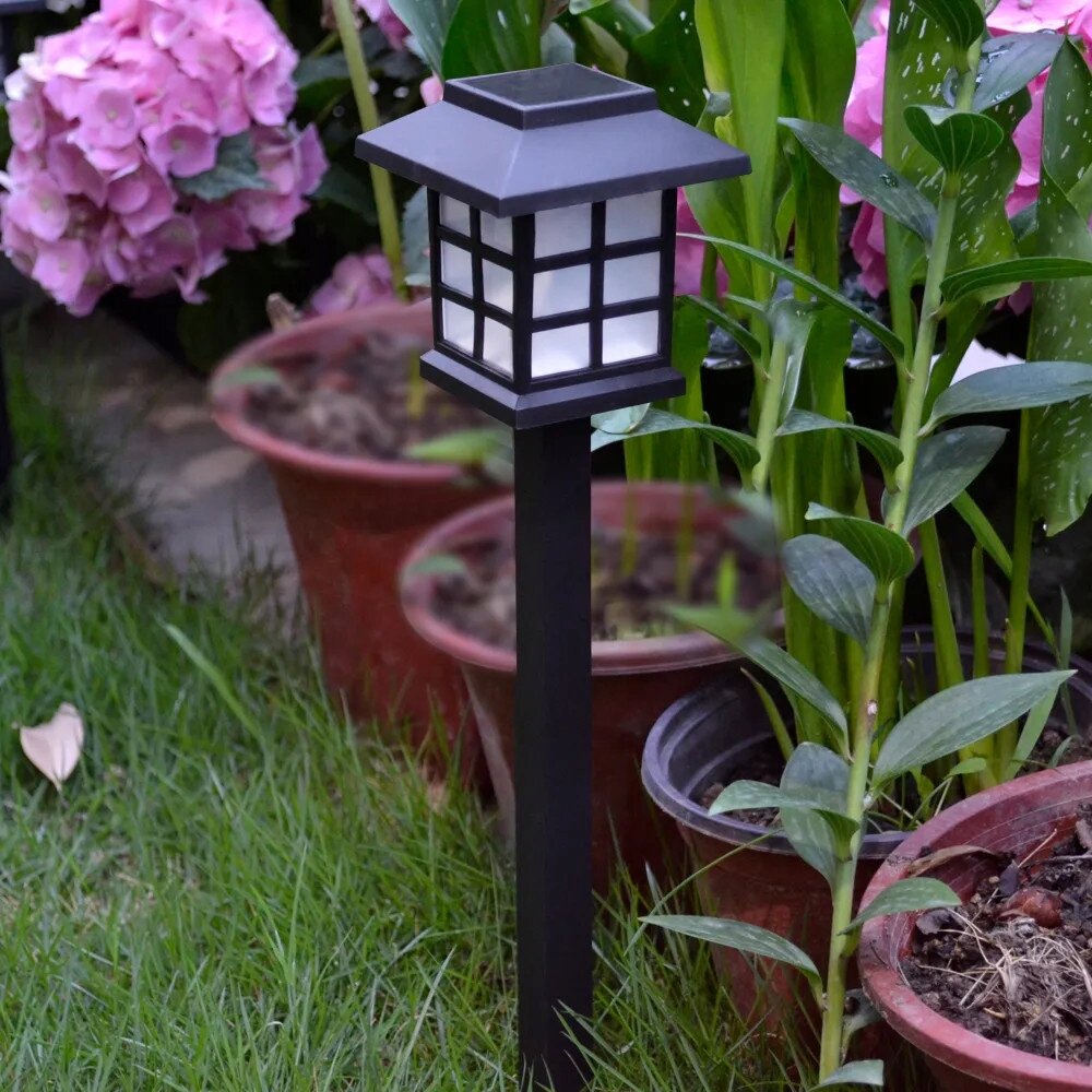 Czarna lampa solarna ogrodowa w kształcie słupka z kwadratowym kloszem, umieszczona na trawniku obok donic z roślinami.