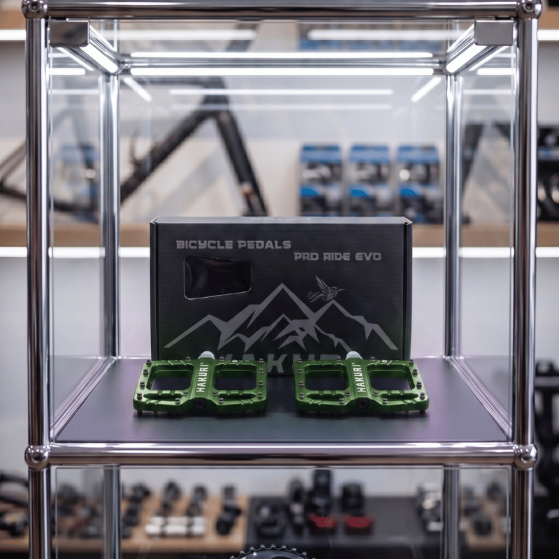 W szklanej gablocie znajdują się ciemnozielone pedały rowerowe z pinami oraz pudełko z napisem \'BICYCLE PEDALS PRO RIDE EVO\'. Na pedałach widoczny jest napis \'HAKURI\'.