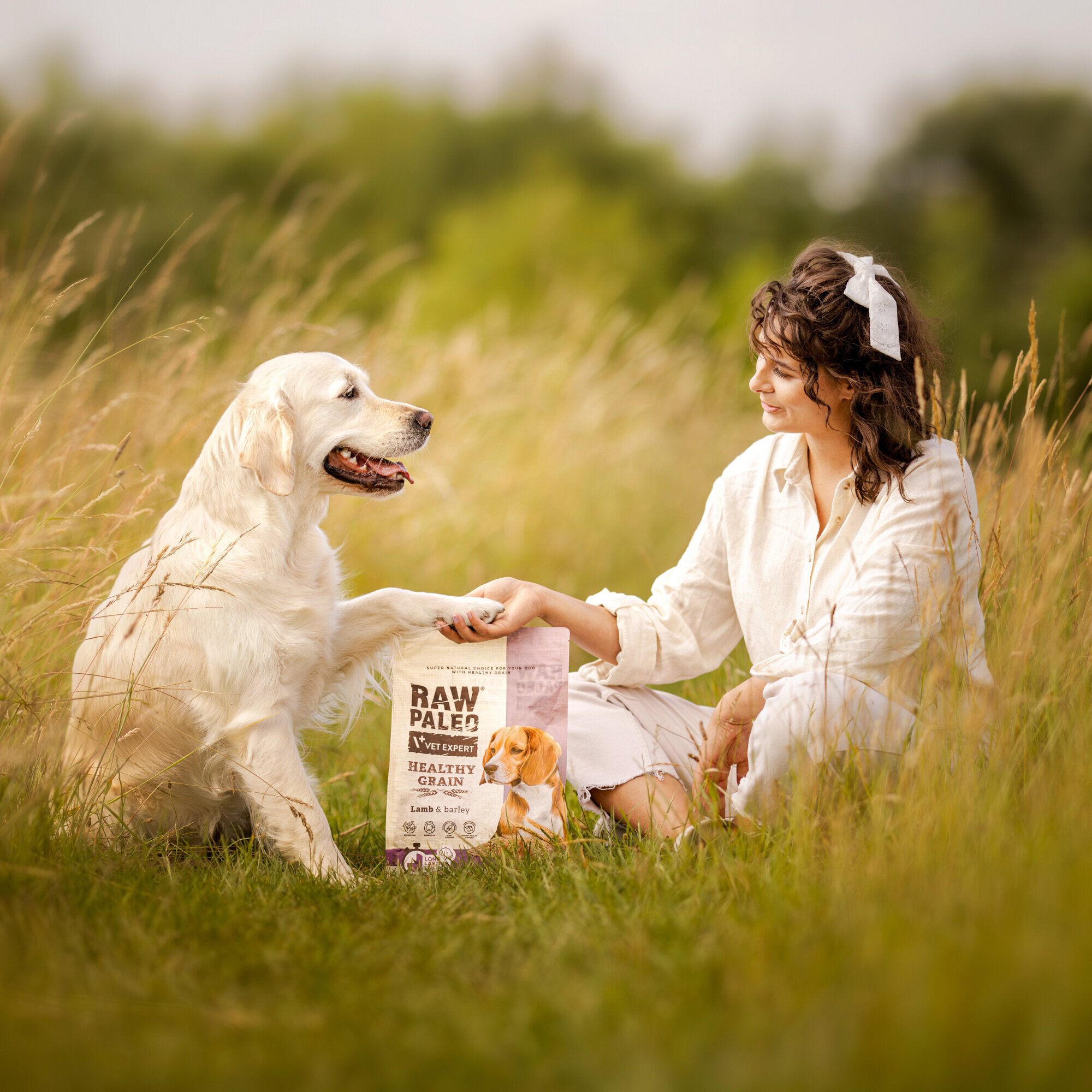 Na obrazie widać psa i osobę siedzącą na trawie obok opakowania karmy dla psów z napisem \'RAW PALEO VET EXPERT HEALTHY GRAIN Lamb & Barley\'. W tle znajduje się zielony krajobraz.