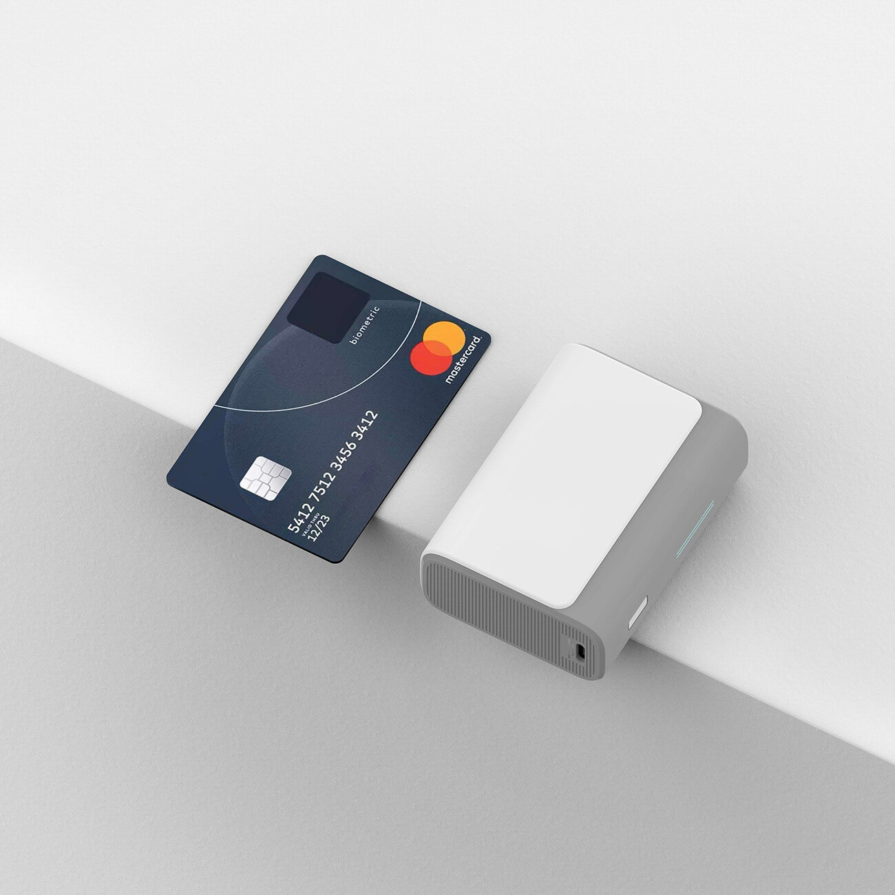 Na obrazie widoczny jest szary power bank oraz karta płatnicza z logo Mastercard. Power bank ma prostokątny kształt z zaokrąglonymi krawędziami.