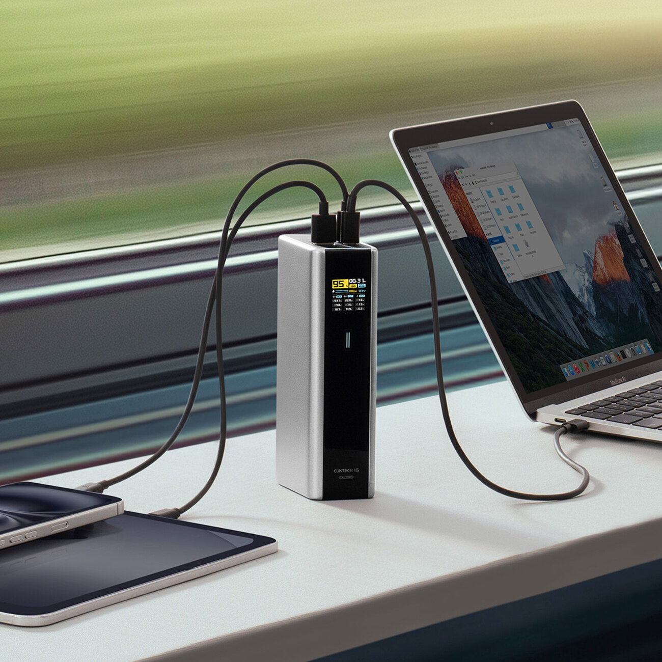 Na obrazie widać szary powerbank z wyświetlaczem, do którego podłączone są dwa urządzenia: laptop i smartfon. Powerbank stoi na stole przy oknie, a na ekranie laptopa widoczne są ikony folderów.