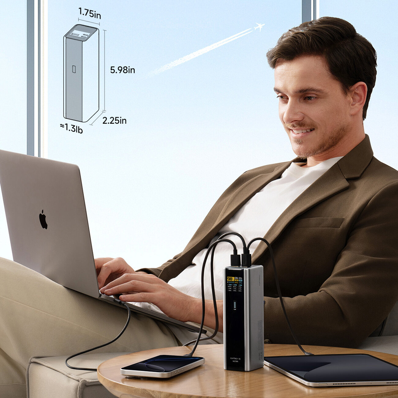 Mężczyzna siedzi na kanapie z laptopem na kolanach, obok niego na stoliku znajduje się powerbank z podłączonymi kablami do urządzeń. Na obrazie widoczne są wymiary powerbanka: 1.75in x 5.98in x 2.25in, waga około 1.3lb, oraz samolot na niebie za oknem.