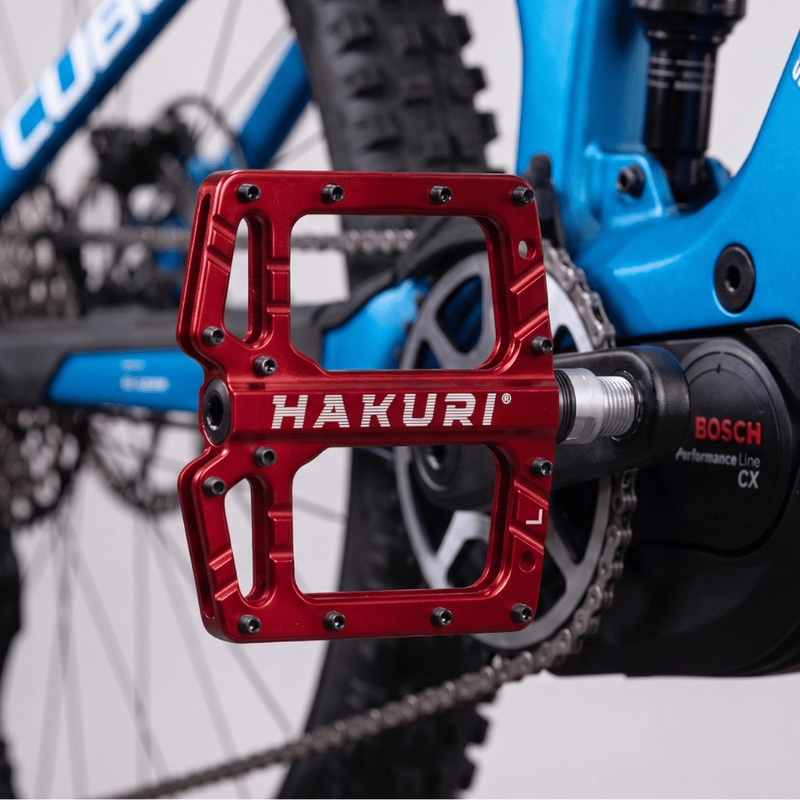 Czerwony pedał rowerowy z napisem \'HAKURI\' zamontowany na niebieskim rowerze. Widoczne jest również logo \'BOSCH Performance Line CX\' na elemencie roweru.