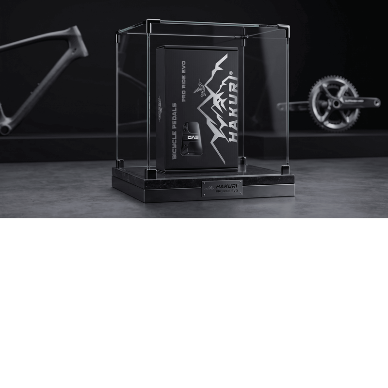 Czarna skrzynka z napisem \'HAKURI\' i \'BICYCLE PEDALS PRO RIDE EVO\' znajduje się w szklanej gablocie na marmurowej podstawie. W tle widoczne są części rowerowe, takie jak rama i korba.