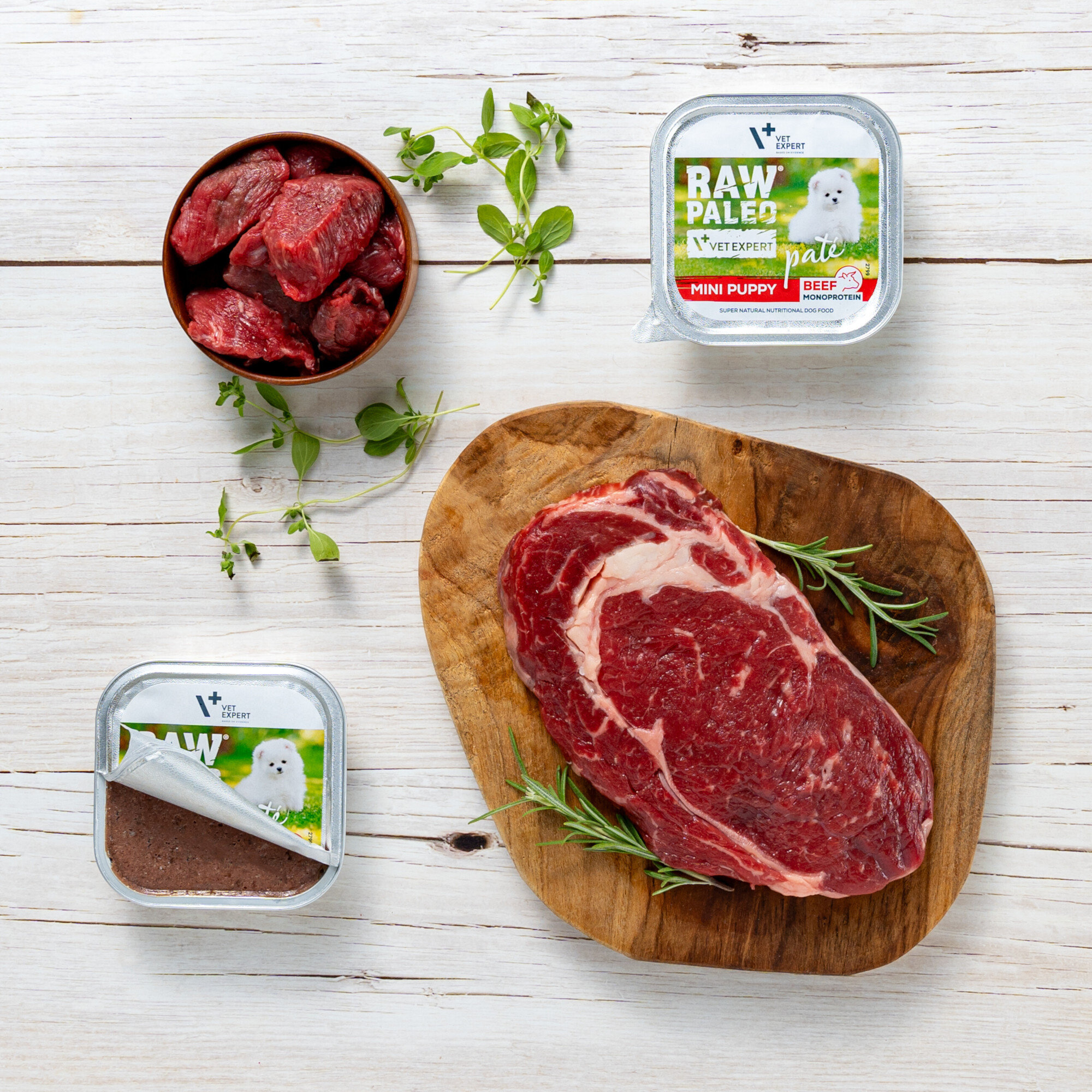Na drewnianej desce leży kawałek surowego mięsa, obok znajdują się zioła. Widać również dwa opakowania produktu \'RAW PALEO VETEXPERT MINI PUPPY BEEF\' oraz miseczkę z kawałkami mięsa.