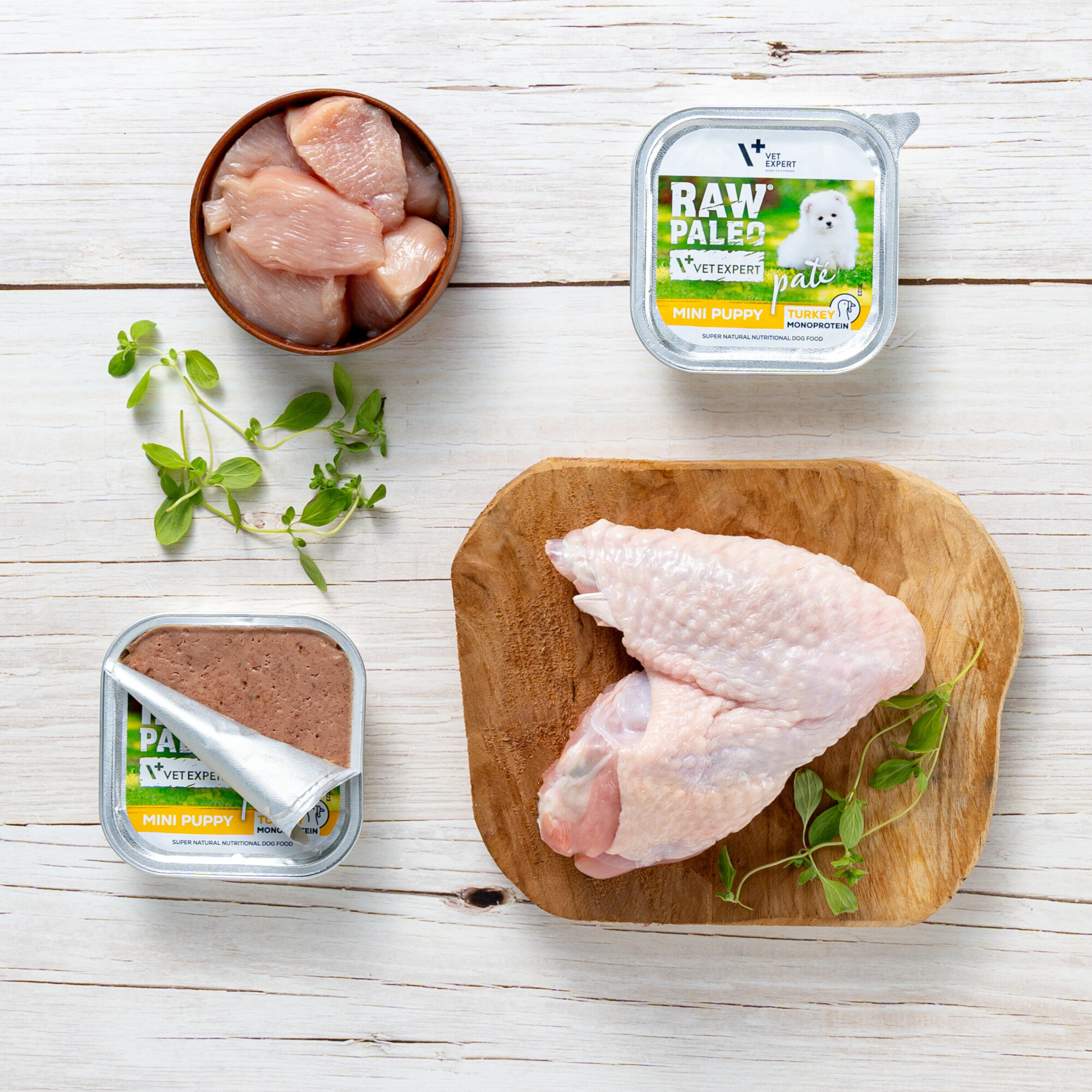 Na drewnianym tle znajduje się miska z kawałkami surowego mięsa, tacka z surowym skrzydłem kurczaka oraz dwa opakowania mokrej karmy dla szczeniąt z etykietą \'RAW PALEO MINI PUPPY TURKEY\'.