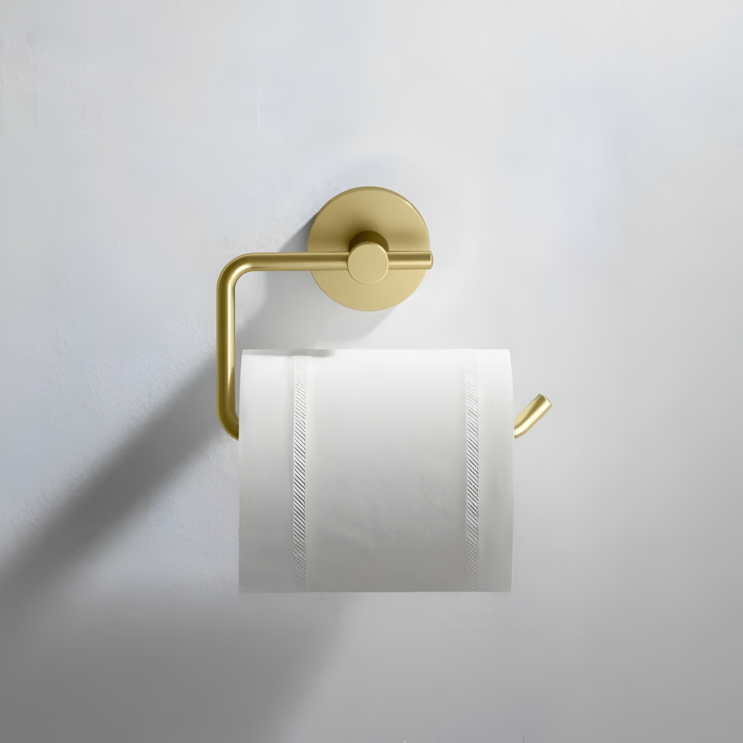 Złoty, metalowy uchwyt na papier toaletowy zamontowany na ścianie, z rolką papieru toaletowego. Uchwyt ma nowoczesny design z okrągłą podstawą i prostym ramieniem.