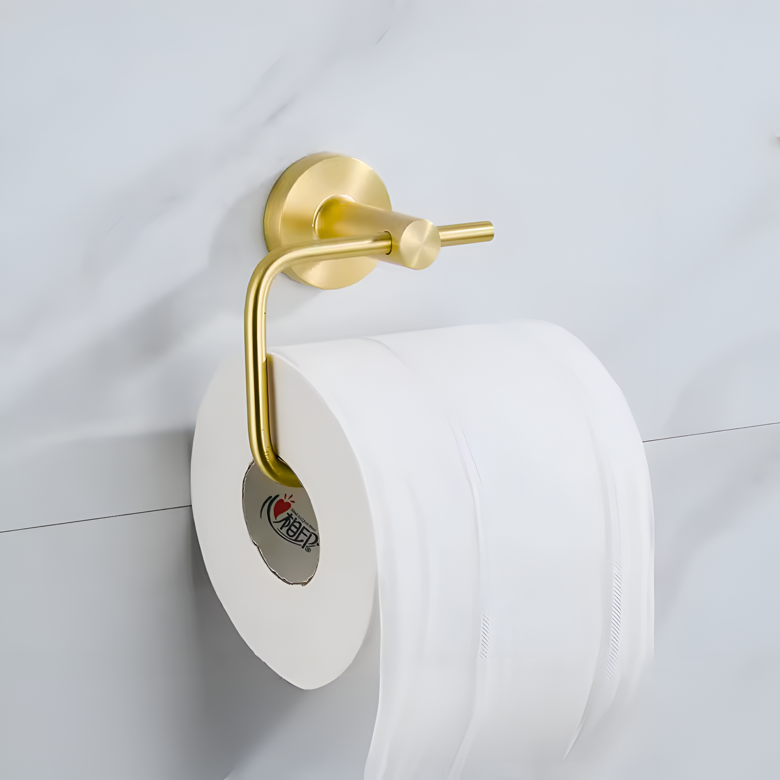 Złoty metalowy uchwyt na papier toaletowy zamontowany na ścianie, z rolką białego papieru toaletowego. Na rolce widoczne jest logo z czerwonym sercem i czarnym napisem w obcym języku.