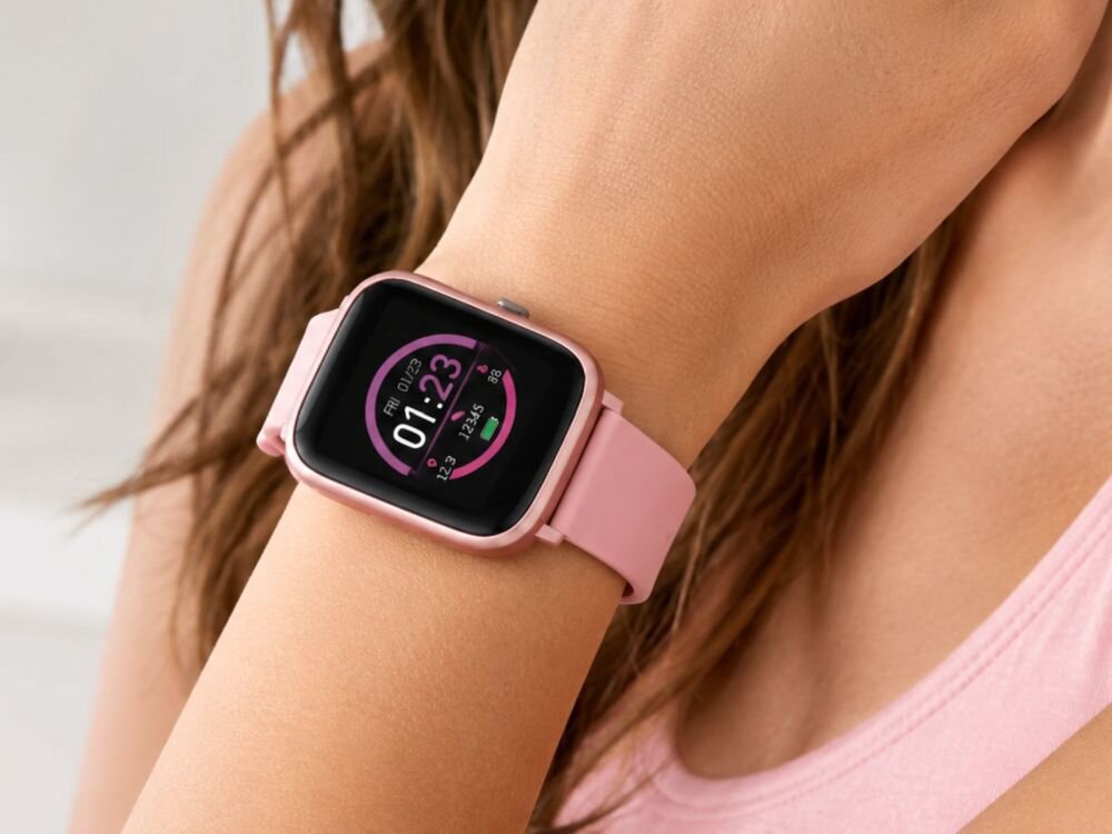 Na zdjęciu widoczny jest różowy smartwatch na nadgarstku osoby, z wyświetlaczem pokazującym godzinę 01:23 oraz datę piątek 01/29. Pasek zegarka jest również różowy, a tło jest rozmyte.