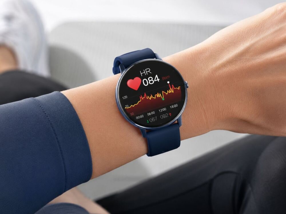 Osoba nosi smartwatch z niebieskim paskiem, na ekranie widoczny jest pomiar tętna z wartością \'HR 084 bpm\' oraz wykres. Tło jest rozmyte, co sugeruje, że osoba siedzi na jasnym fotelu.