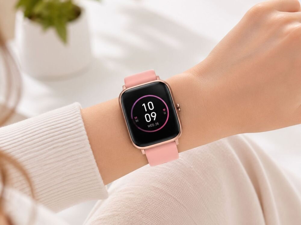 Na zdjęciu widoczny jest smartwatch z różowym paskiem na nadgarstku. Na ekranie zegarka wyświetla się godzina 10:09 oraz dzień tygodnia \'WED 28\'.