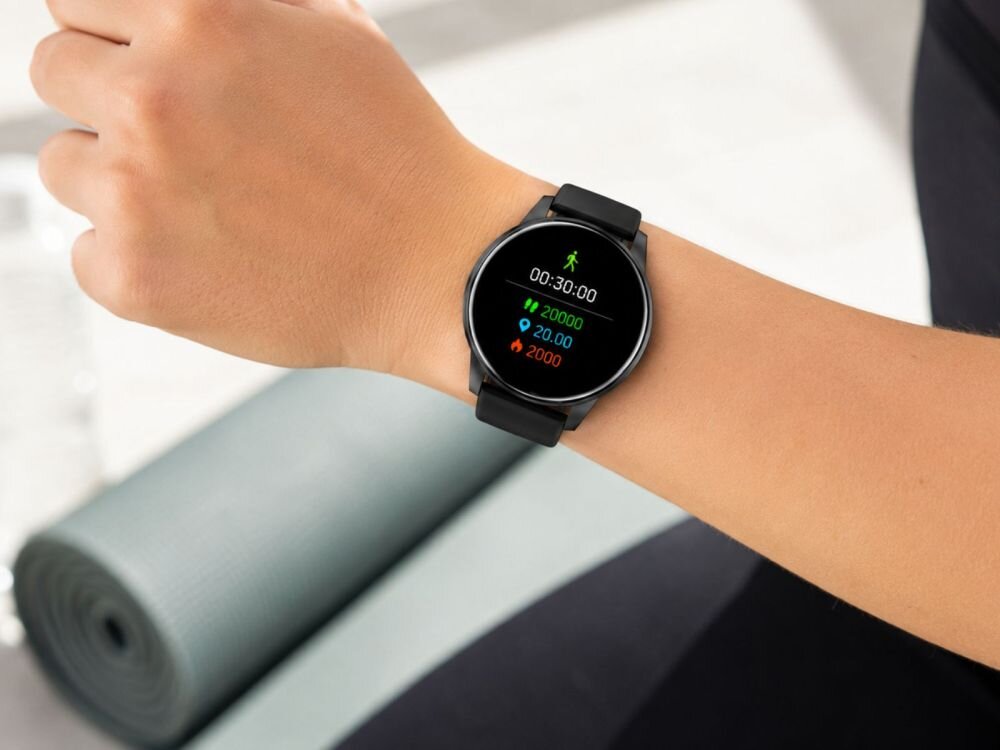 Osoba nosi czarny smartwatch z okrągłym wyświetlaczem, na którym widoczny jest czas 00:30:00 oraz liczby 20000, 20.00 i 2000 w różnych kolorach. W tle znajduje się zwinięta mata do ćwiczeń.