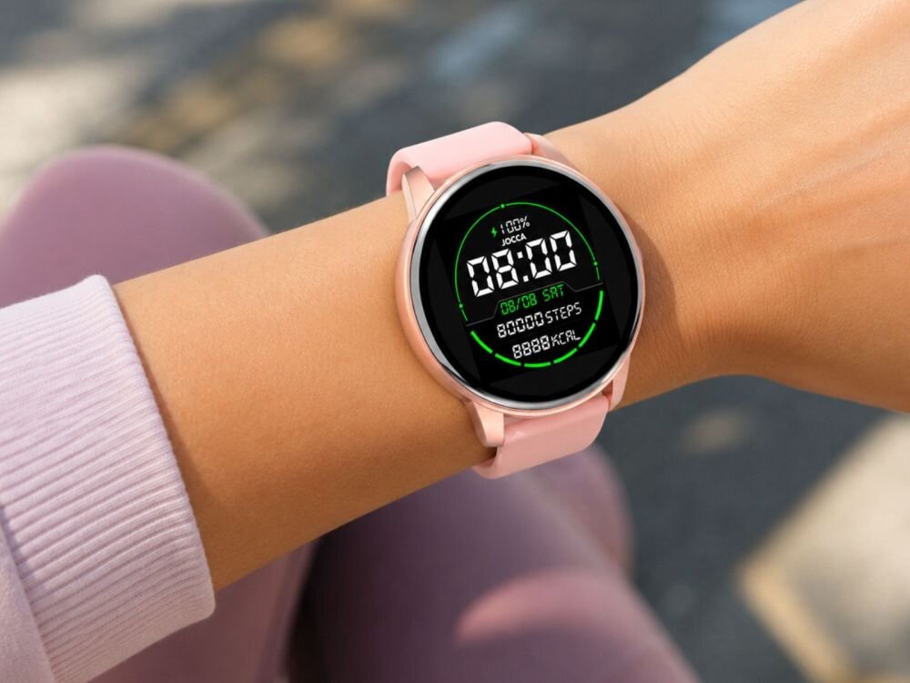 Osoba nosi różowy smartwatch z okrągłym wyświetlaczem, na którym widoczny jest tekst: \'JOCCA\', \'08:00\', \'80,000 STEPS\', \'8888 KCAL\'.