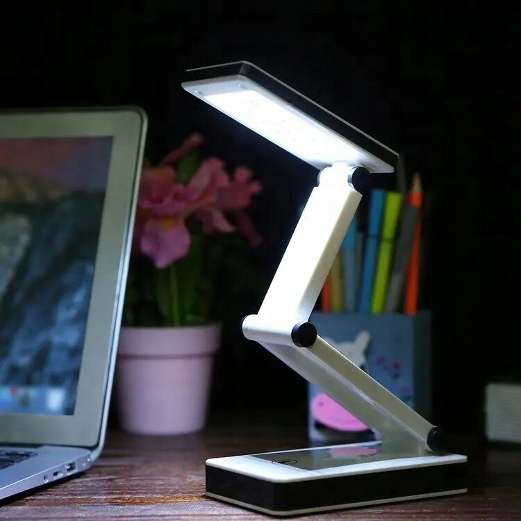 Na biurku znajduje się biała, składana lampka LED o nowoczesnym designie, obok laptopa i doniczki z różowymi kwiatami. W tle widoczne są kolorowe przybory do pisania w pojemniku.