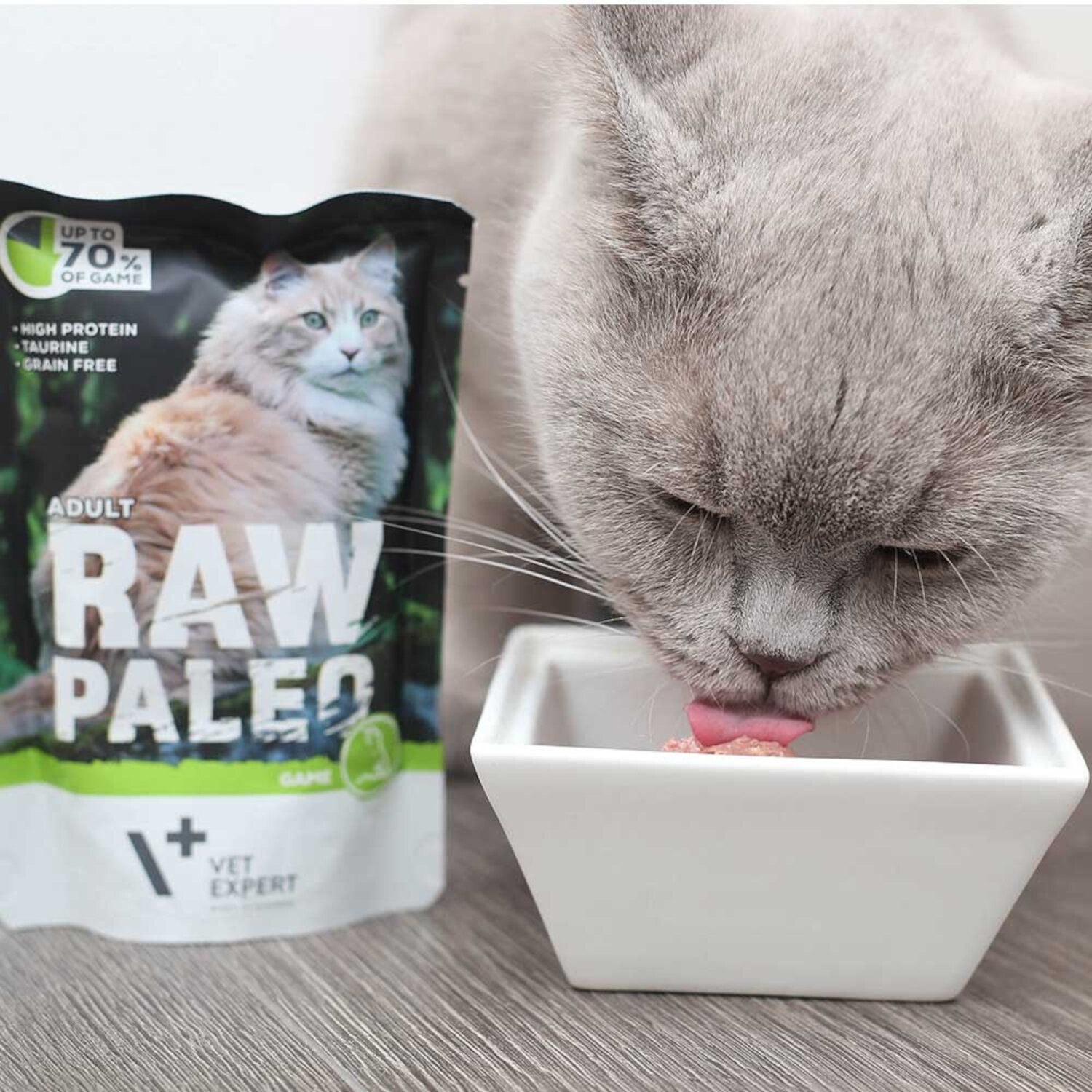 Na zdjęciu widać szarego kota jedzącego z białej miski oraz opakowanie karmy RAW PALEO z wizerunkiem kota i napisami: \'UP TO 70% OF GAME\', \'HIGH PROTEIN\', \'TAURINE\', \'GRAIN FREE\', \'ADULT RAW PALEO\', \'VET EXPERT\'.