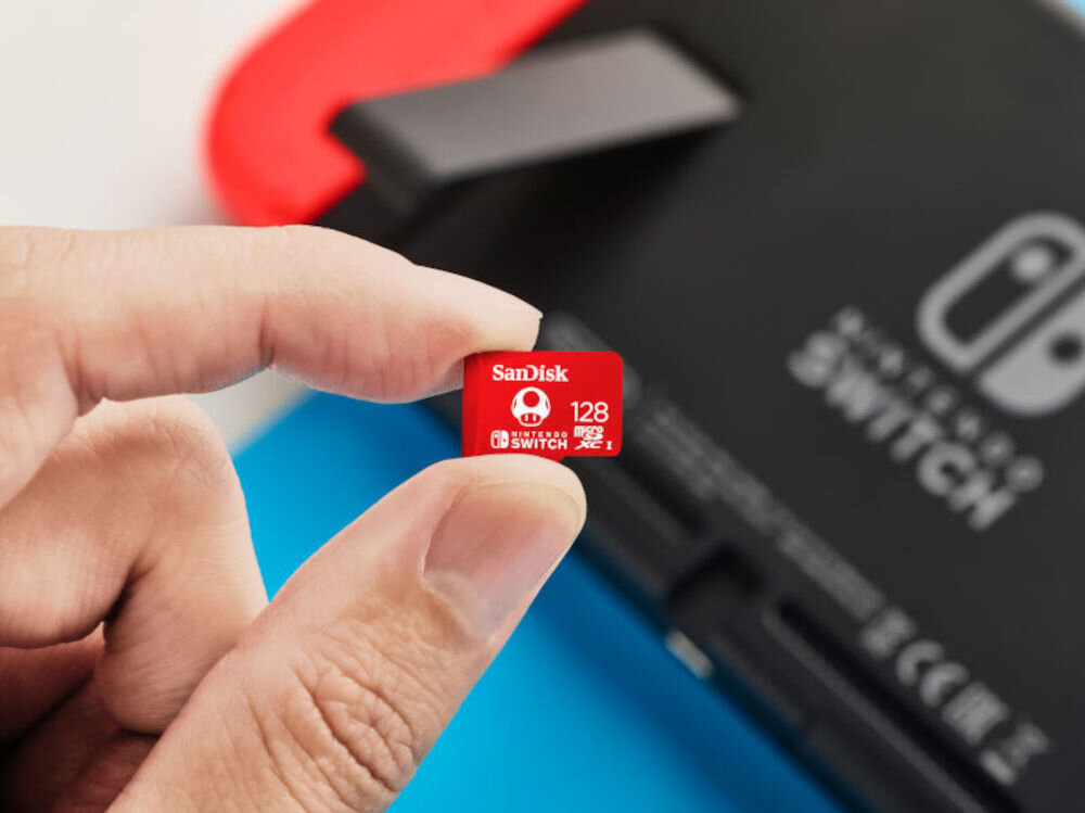 Osoba trzyma czerwoną kartę pamięci z napisem \'SanDisk 128 GB Nintendo Switch\'. W tle widoczna jest część urządzenia z logo Nintendo Switch.