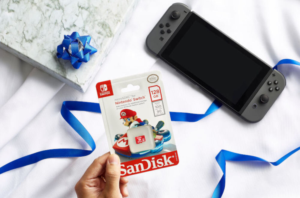 Osoba trzyma opakowanie karty pamięci SANDISK microSDXC 128GB do Nintendo Switch z grafiką postaci z gry. Obok leży konsola do gier i niebieska wstążka na białym tle.
