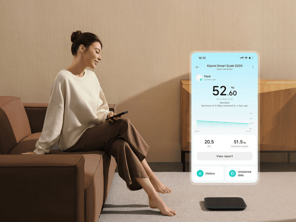 Osoba siedzi na kanapie z telefonem w ręku, a przed nią na podłodze znajduje się waga łazienkowa. Na ekranie telefonu widoczny jest interfejs aplikacji \'Xiaomi Smart Scale S200\' z informacjami o wadze użytkownika.