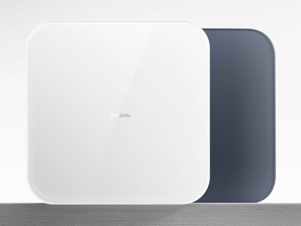 Na obrazie widoczna jest biała waga łazienkowa z zaokrąglonymi rogami, z napisem \'xiaomi\' na środku. W tle znajduje się ciemniejszy element o podobnym kształcie.