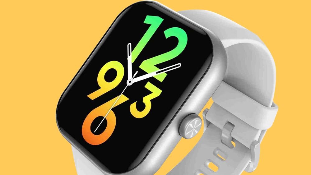 Smartwatch z prostokątnym wyświetlaczem, na którym widoczny jest kolorowy zegar z dużymi cyframi 12, 3, 6 i 9. Pasek zegarka jest jasny, a obudowa ma metaliczny wygląd.